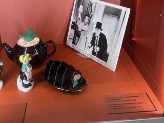 Questa immagine mostra una vetrina espositiva dal fondo arancione contenente oggetti da collezione legati al mondo dei fumetti e del cinema d'epoca. Sulla sinistra è presente un set da tè di Andy Capp (Wade, 1997), composto da: una teiera nera il cui coperchio riproduce la testa del celebre personaggio con il suo tipico berretto a scacchi verde. Due piccole statuine in ceramica dei protagonisti, Andy e sua moglie Flo. Un portatovaglioli nero decorato con un'illustrazione dei due personaggi. Sulla destra si trova una fotografia in bianco e nero poggiata verticalmente, che ritrae una scena del film Jiggs and Maggie in Jackpot Jitters (1949). La foto mostra un uomo con cappello a cilindro e una donna che regge un oggetto alto. In basso a destra, un cartellino esplicativo bilingue (italiano e inglese) fornisce i dettagli storici sulla foto di scena e sul regista William Beaudine.