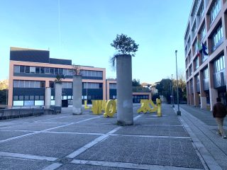 "Una veduta di una piazza urbana pavimentata, caratterizzata da un'architettura moderna. Al centro, pilastri cilindrici in cemento fungono da base per piccoli alberi posizionati sulla sommità, creando un elemento di design urbano. Accanto a questi, sculture metalliche di grandi dimensioni di colore giallo vivace, che richiamano forme astratte o lettere, sono disposte sul pavimento in pietra. Sulla sinistra si trova un edificio a più piani con una facciata geometrica in toni di marrone e grigio, mentre sulla destra si staglia una struttura più alta con ampie vetrate. L'atmosfera è quella di uno spazio pubblico contemporaneo, inquadrato da un cielo sereno e luminoso."