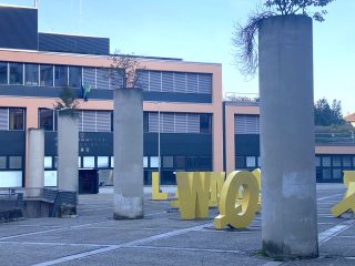"Veduta di una piazza pubblica pavimentata in lastre di pietra e sampietrini, caratterizzata da un'architettura razionalista. In primo piano, tre alti pilastri cilindrici in cemento sostengono dei piccoli alberi sulla sommità. Accanto a essi, alcune sculture metalliche tridimensionali di colore giallo, dalla forma astratta che ricorda caratteri tipografici, sono collocate direttamente sul pavimento. Sullo sfondo, un edificio amministrativo a due piani con facciata in tonalità pesca e ampie finestre a nastro con persiane grigie, su cui si legge l'iscrizione 'INDUSTRIA ADRIATICO'. Il cielo è limpido, a indicare una giornata soleggiata."